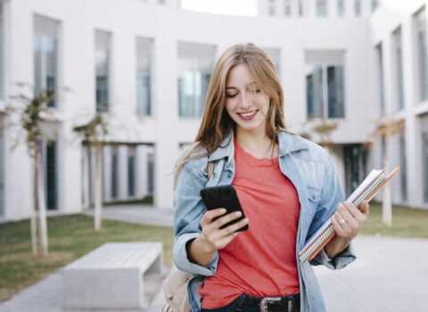 smiling-young-blond-female-student-using-smart-phone-at-university-TCEF01375